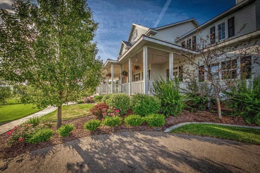 A Tradition Homes farmhouse: a white two-story home with a deep wraparound porch, hanging flower baskets, and landscaped beds along a curving drive